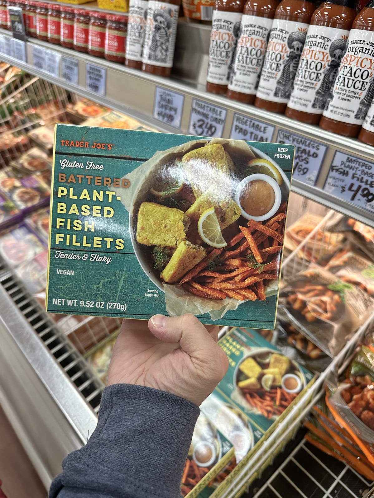 Hand holding Trader Joe's gluten-free battered plant-based fish filets by freezer case.
