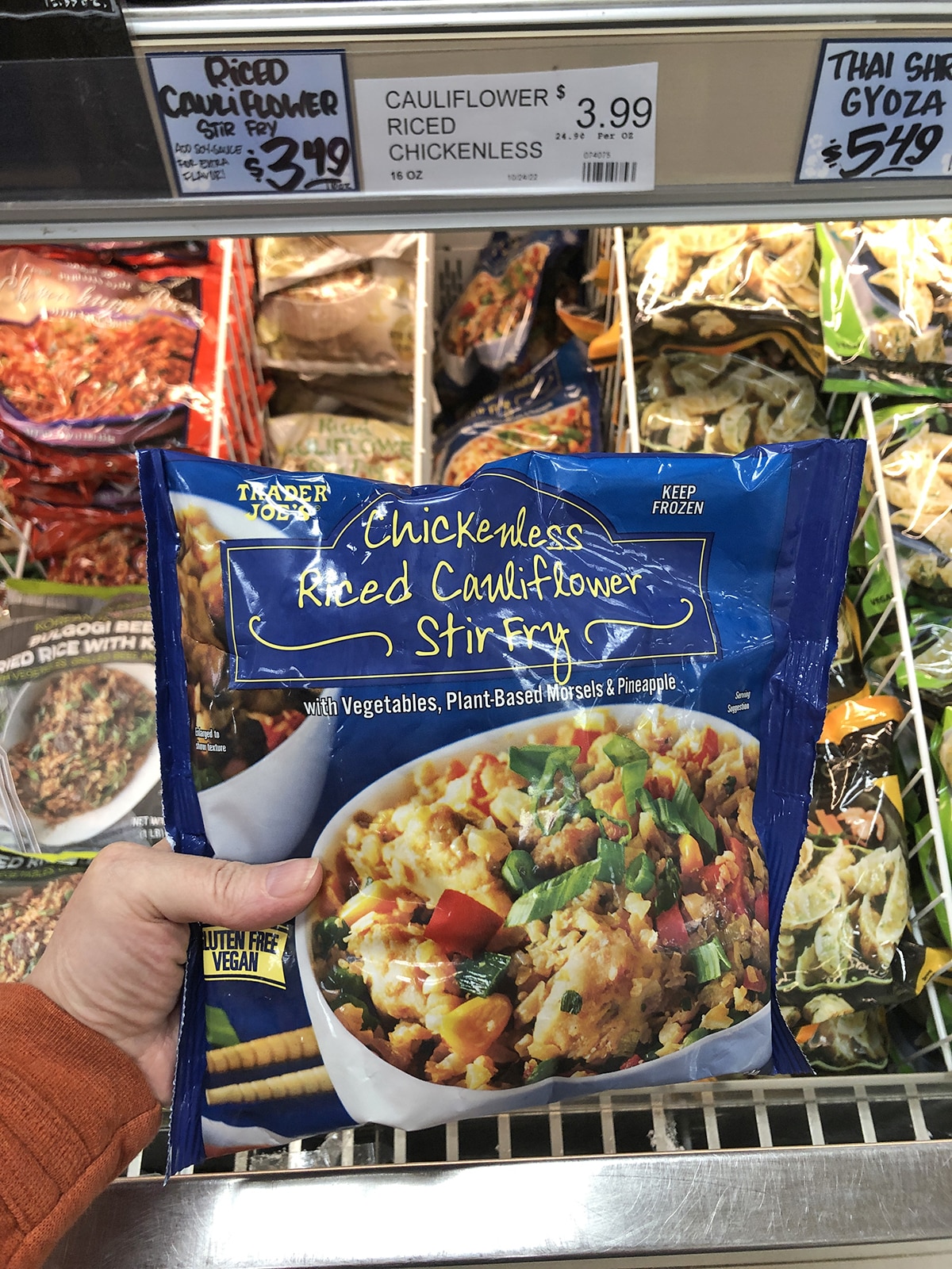 Hand holding chickenless riced cauliflower stir-fry from Trader Joe's in front of frozen counter.