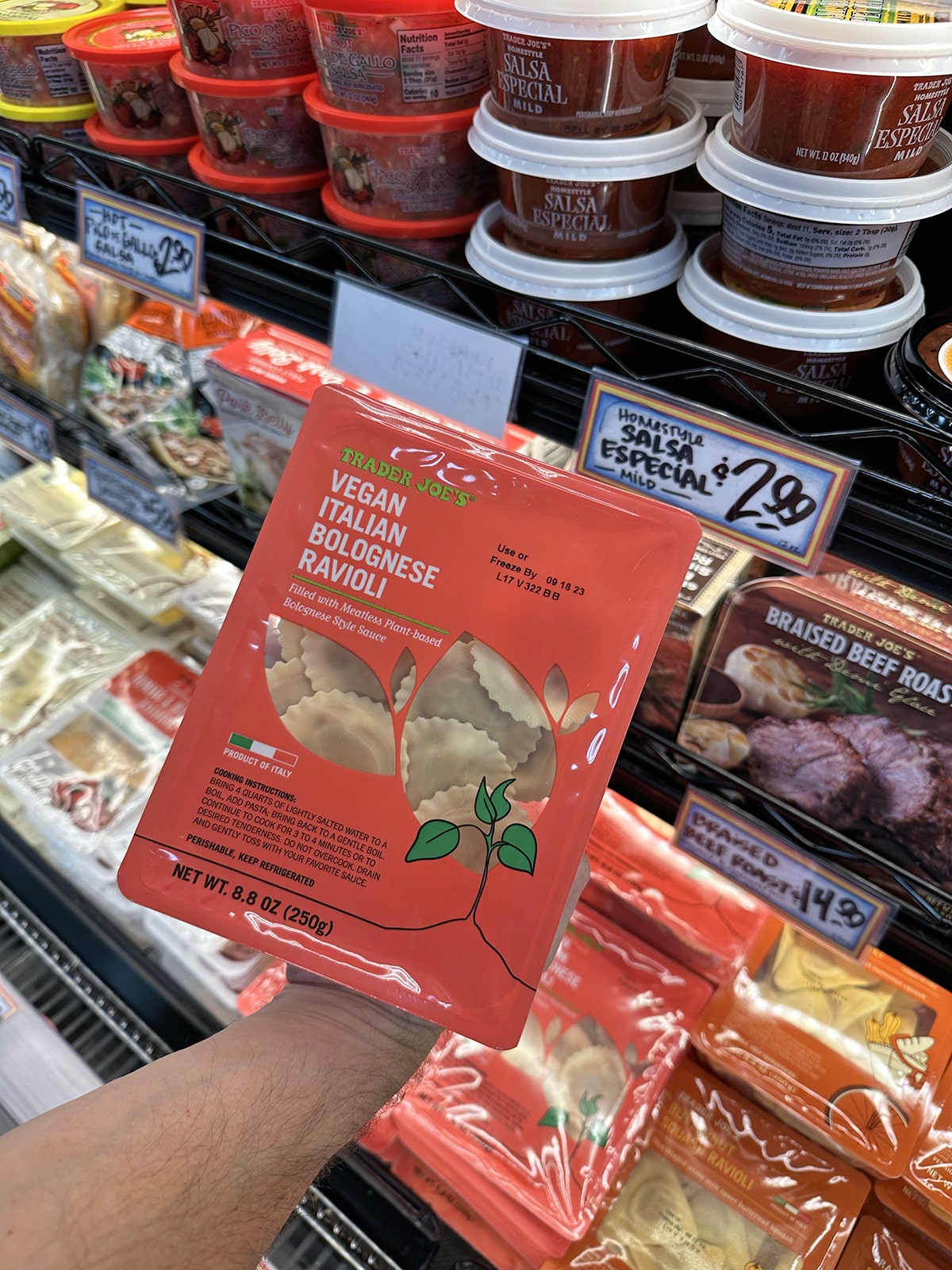 Hand holding Trader Joe's vegan Italian bolognese ravioli in front of refrigerated case.