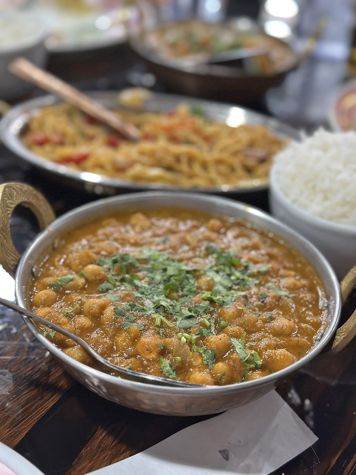 Chana masala in kadhai on table by rice and noodles.