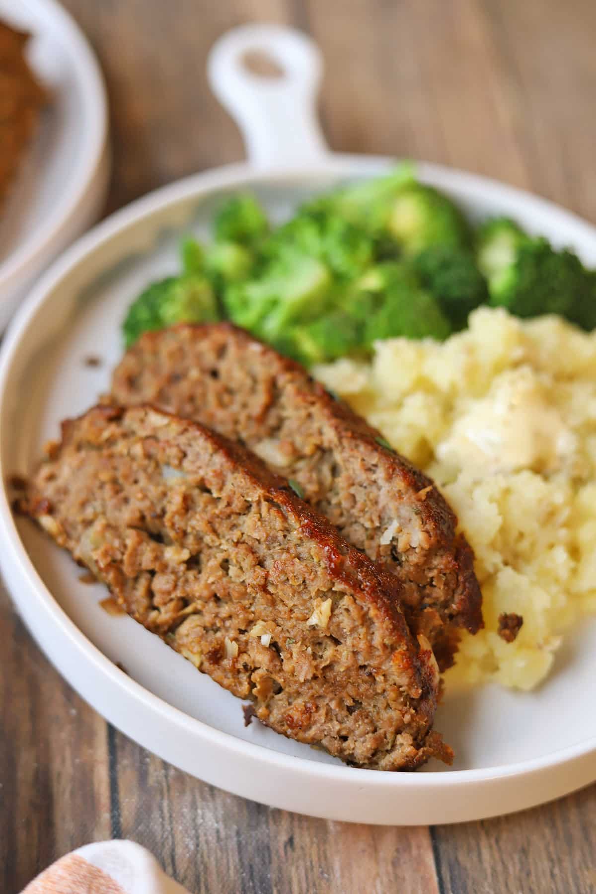 Thick slices of moist vegan meatloaf on plate with mashed potatoes and broccoli.