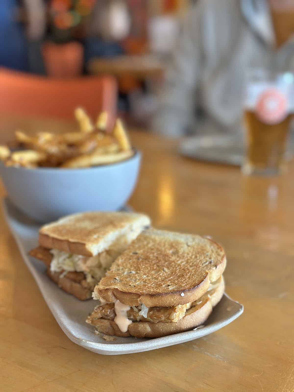 Tempeh reuben on plate by bowl of french fries.