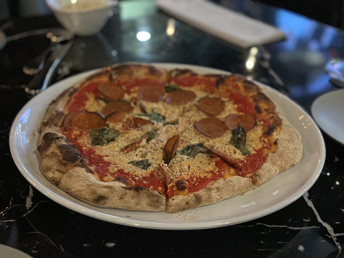 Wood-fired pizza on platter, topped with vegan pepperoni, vegan mozzarella, and basil on marinara.