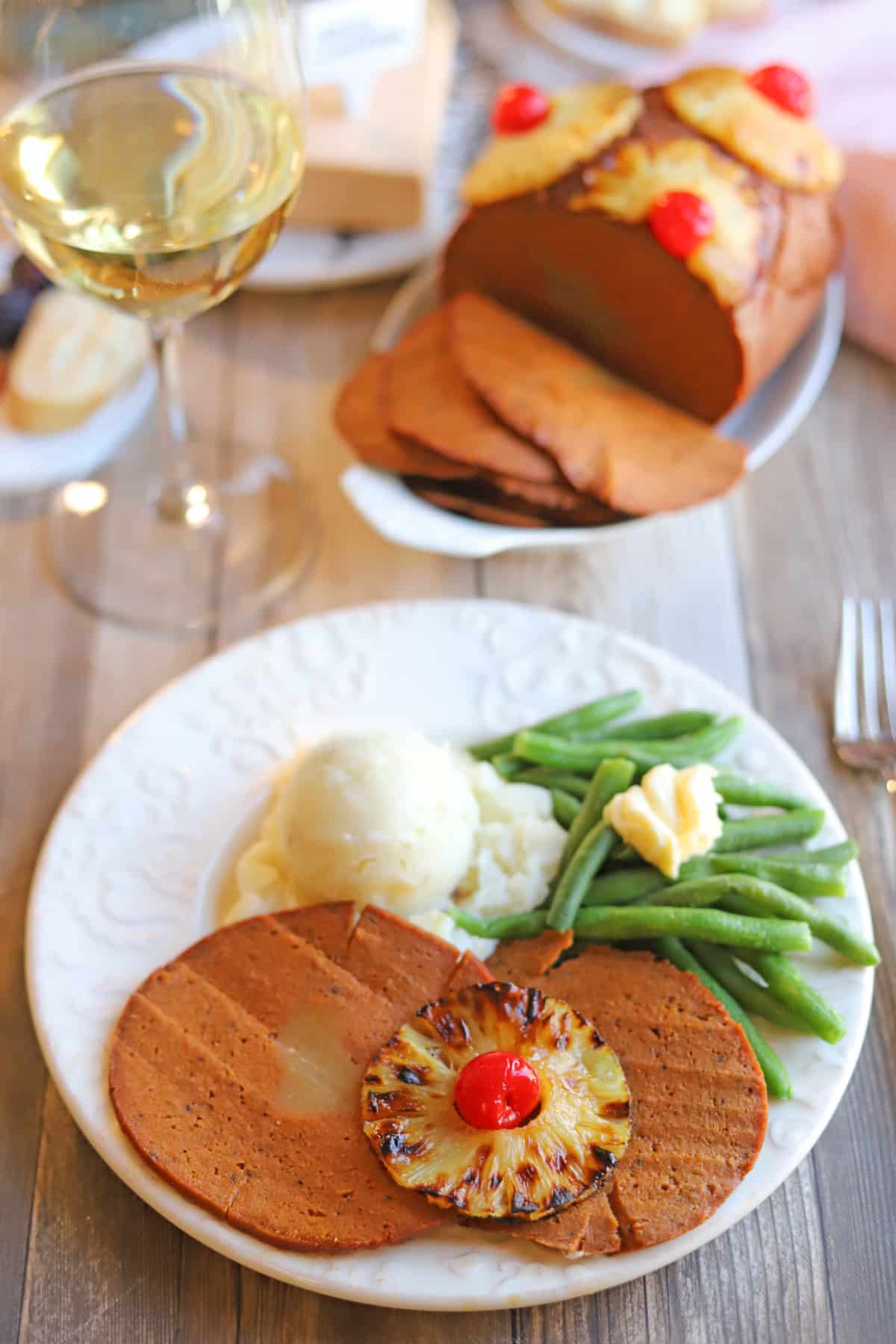 Sliced vegan ham on plate with caramelized pineapple and maraschino cherries, green beans, and mashed potatoes.