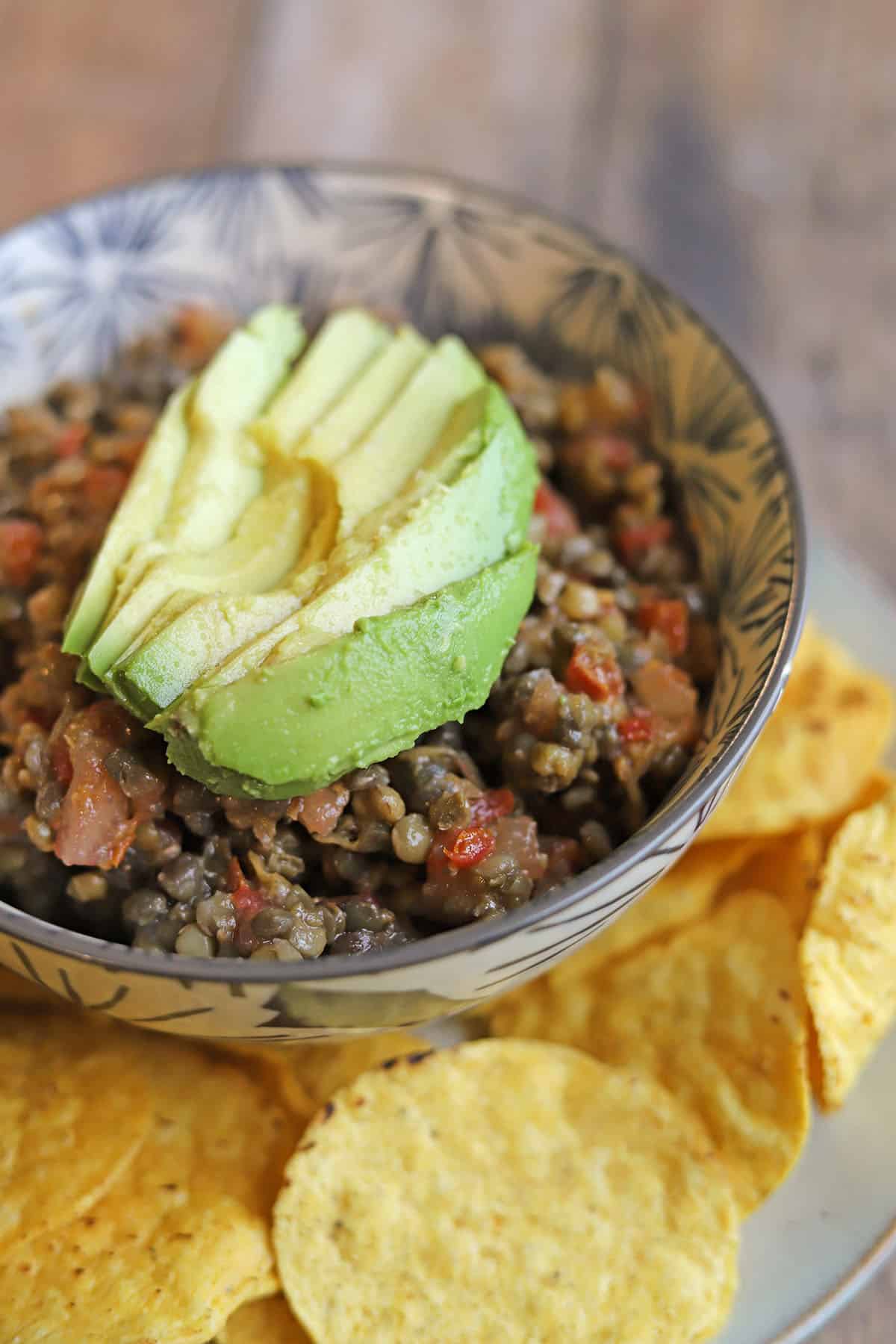 Steamed lentils mixed with bruschetta sauce, topped with sliced avocado in bowl by tortilla chips.