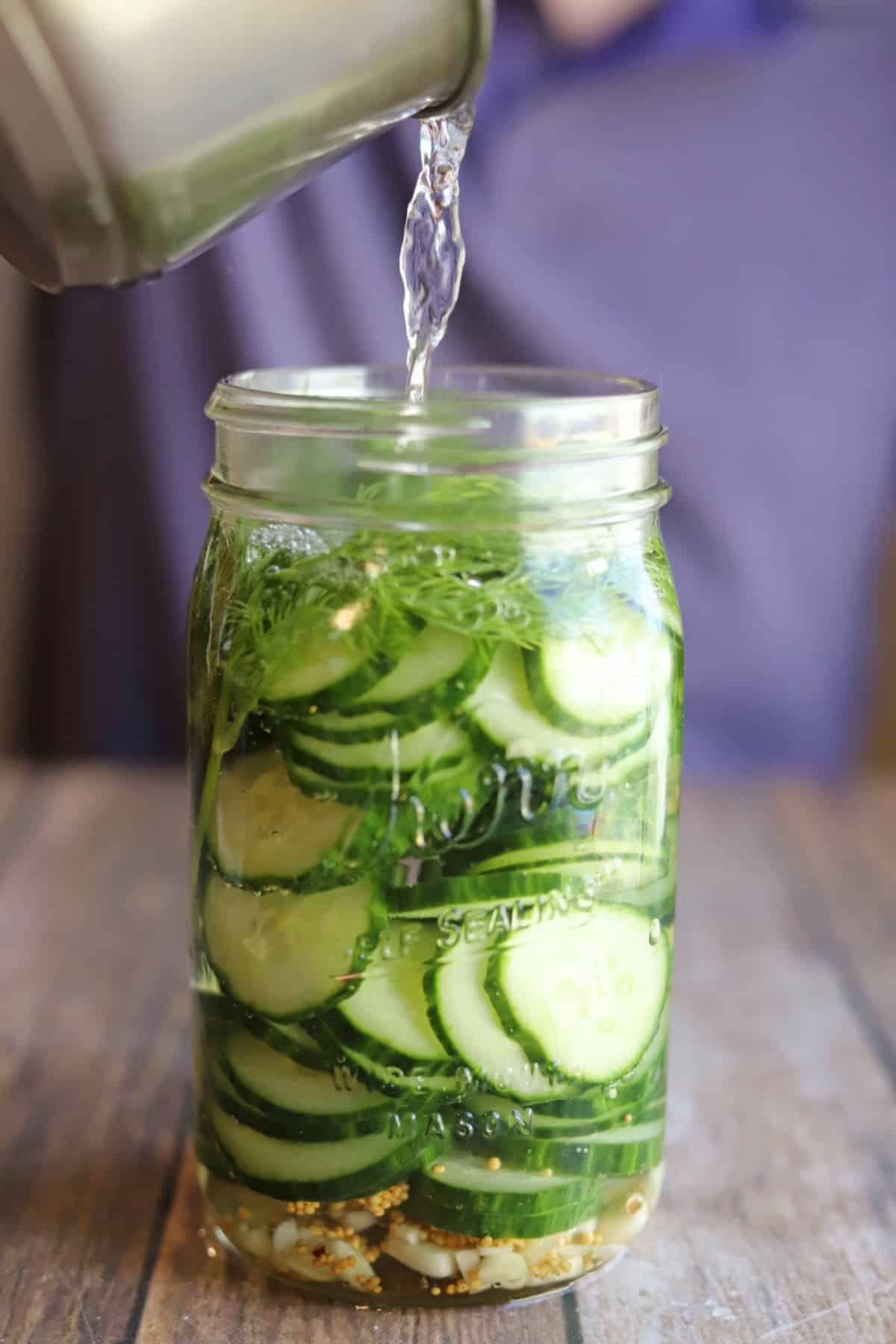 Brine being poured onto thinly sliced cucumber with mustard seeds, garlic, and fresh dill.