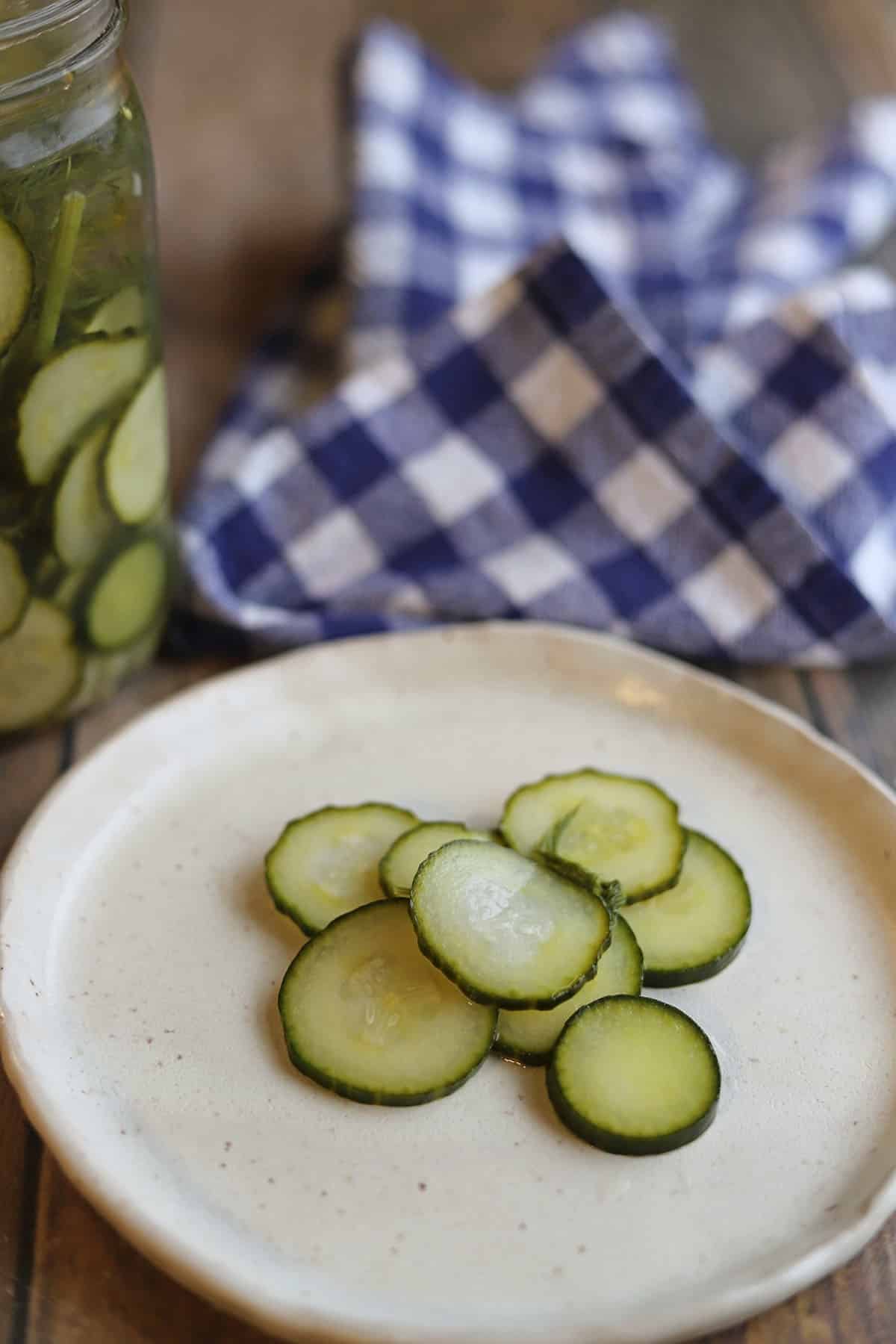 Sliced cucumber refrigerator pickles on a plate by jar and gingham napkin.