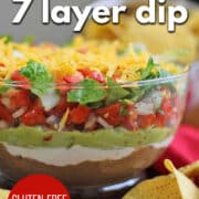 Text overlay: vegan 7 layer dip, gluten-free party dip, cadryskitchen.com. 7-layer dip on table with rows of refried beans, guacamole, vegan sour cream, pico de gallo, and salad.