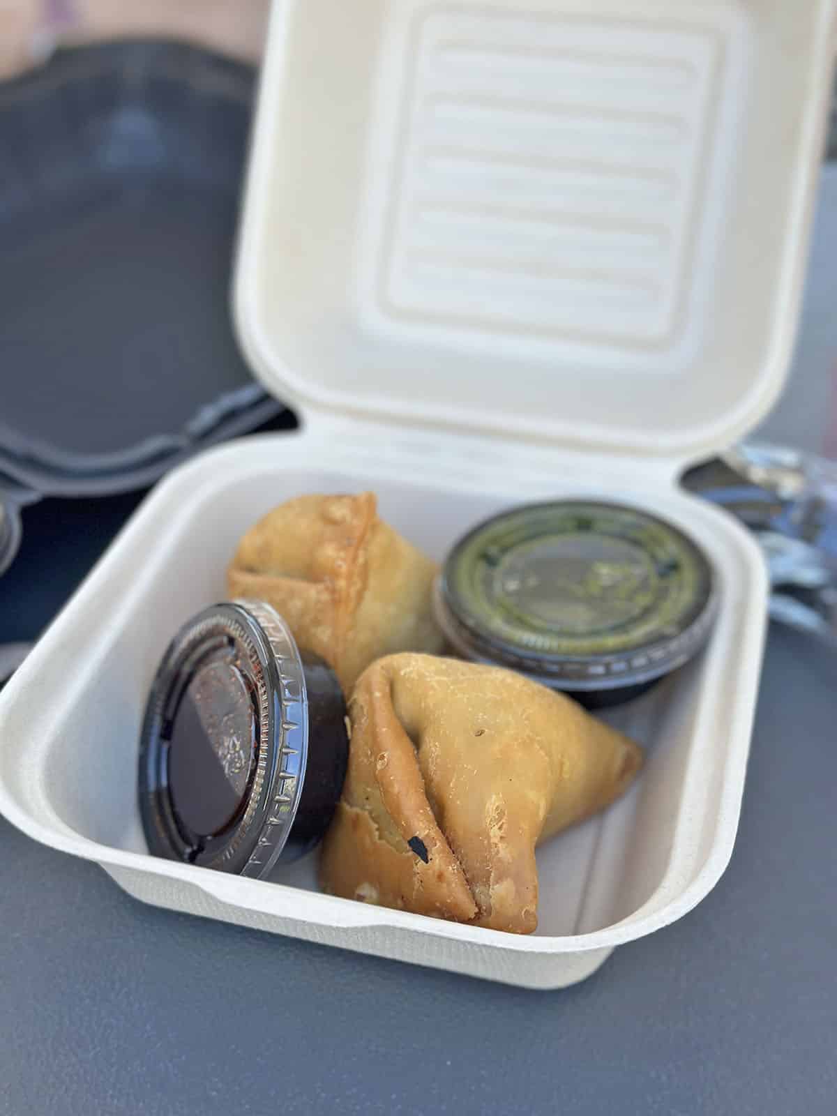 Vegetable samosas in a to-go box with containers of cilantro and tamarind chutneys.
