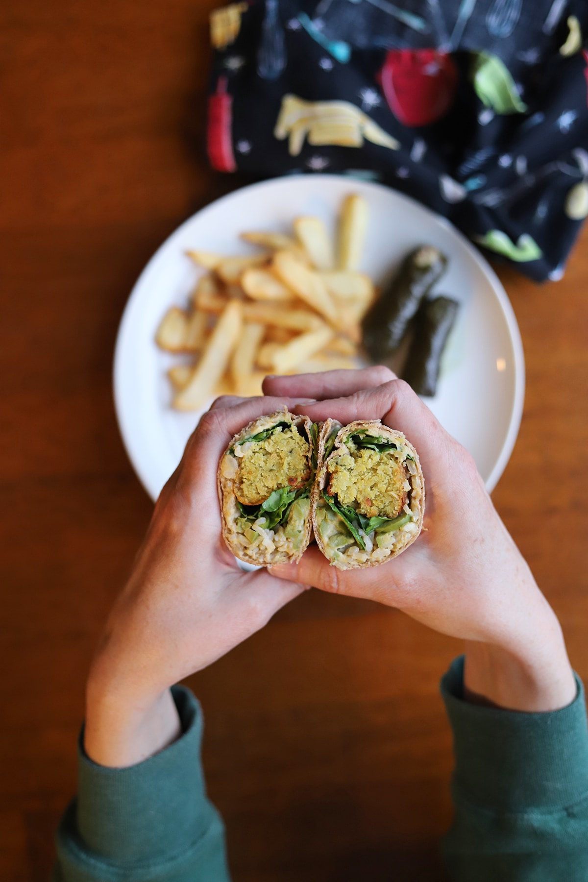 Hands holding falafel wrap over plate with fries and dolmas.