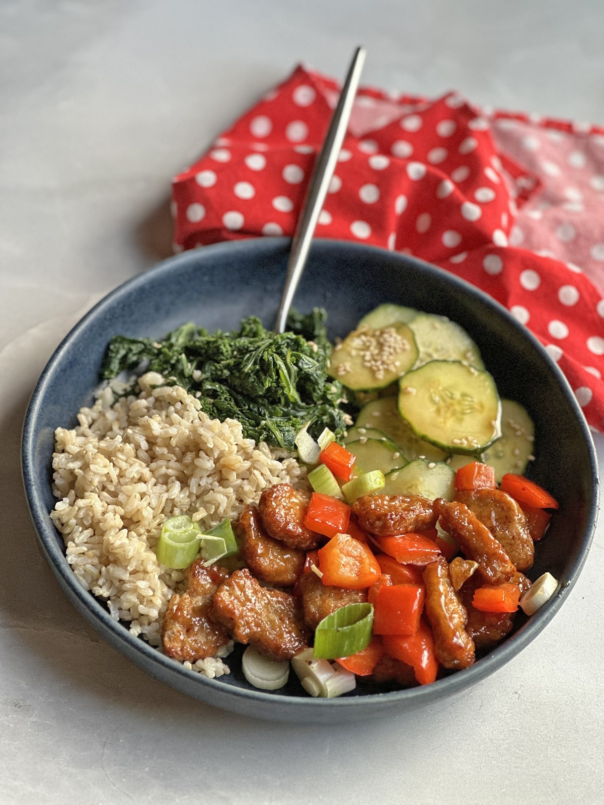 Vegan orange chicken with peppers and onions in bowl with sesame kale, sesame cucumbers, and brown rice.