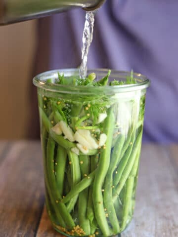 Brine being poured onto green beans with fresh dill and slivered garlic.