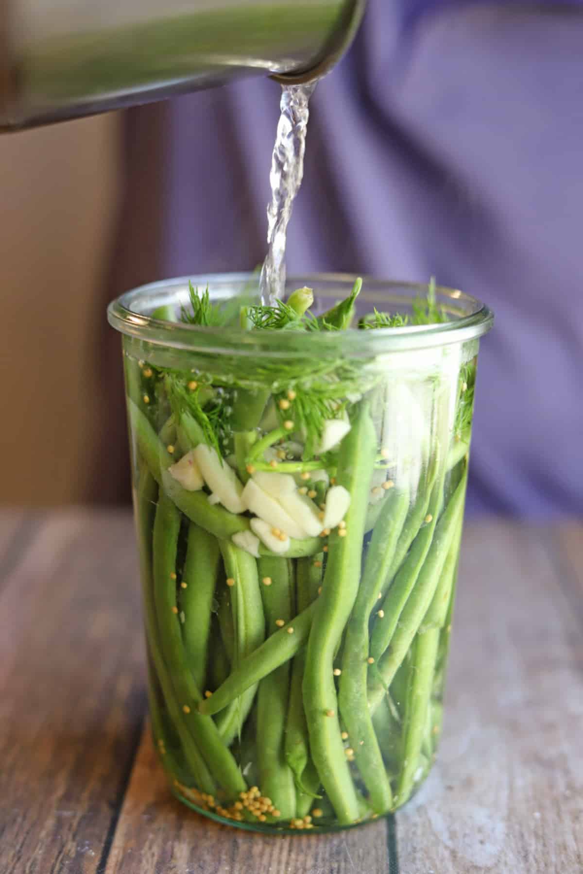 Brine being poured onto green beans with fresh dill and slivered garlic.