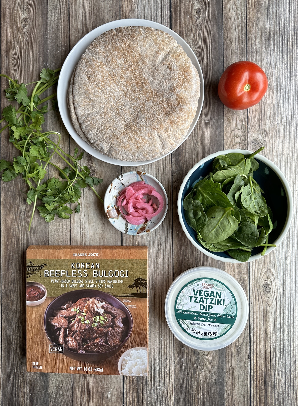 Package of Trader Joe's beefless bulgogi on table with vegan tzatziki, baby spinach, tomato, pickled onions, cilantro, and pita bread.