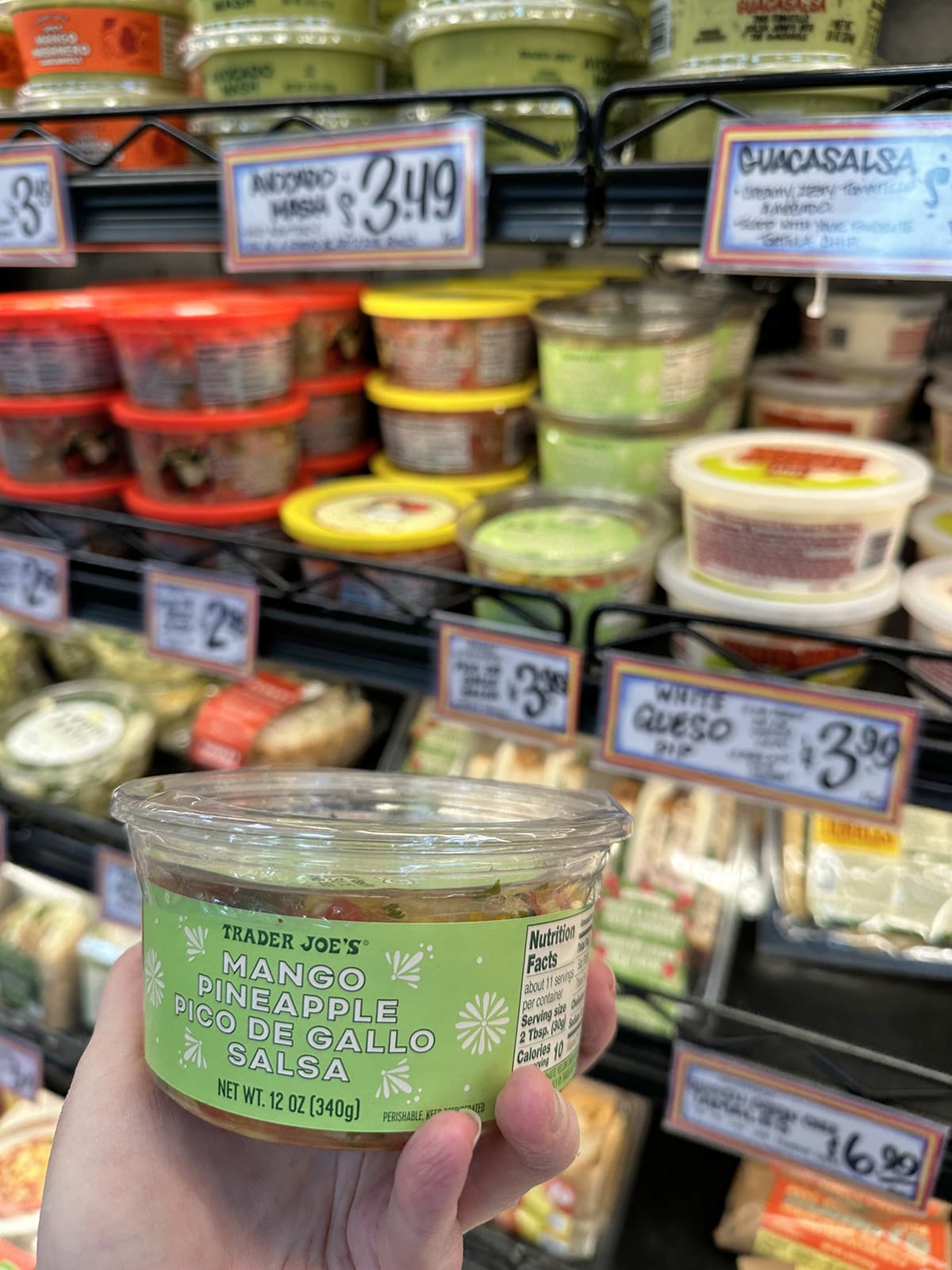 Hand holding container of mango pineapple pico de gallo salsa by refrigerated case at Trader Joe's.