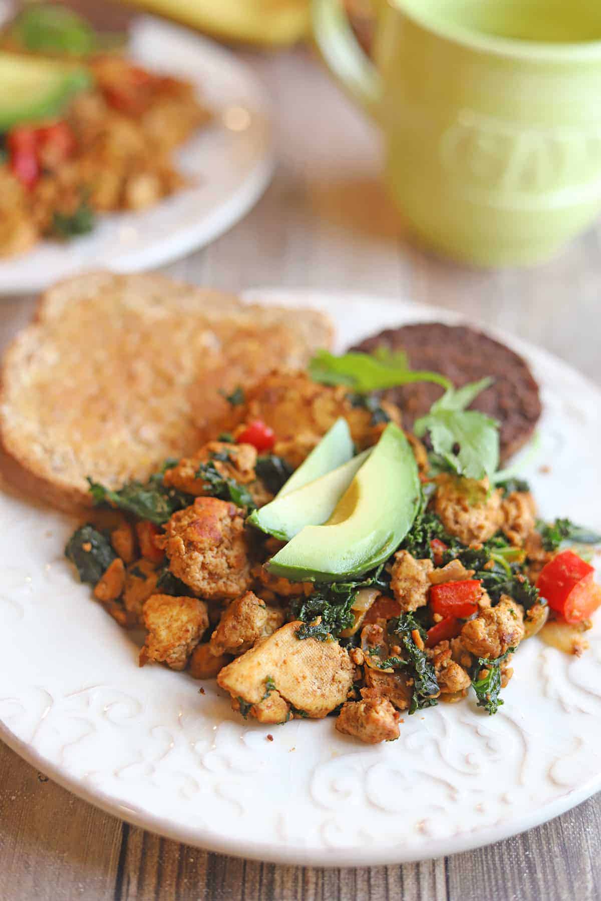 Tofu scramble breakfast with kale and avocado on plate with toast and veggie sausage.