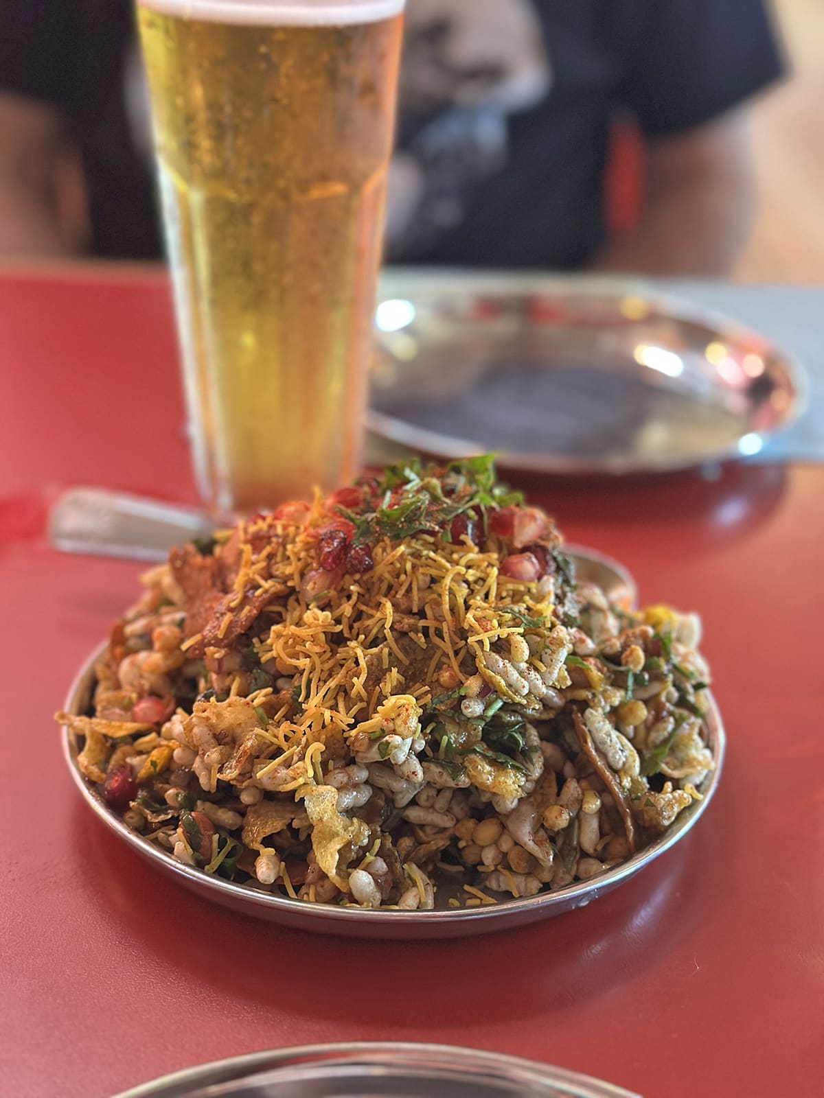 Disco bhel on metal platter with poori, puffed rice, boondi, corn poha, sev, onion, cilantro, roasted peanuts, tamarind chutney, green chutney, bhel mix, and pomegranate by tall glass of beer.