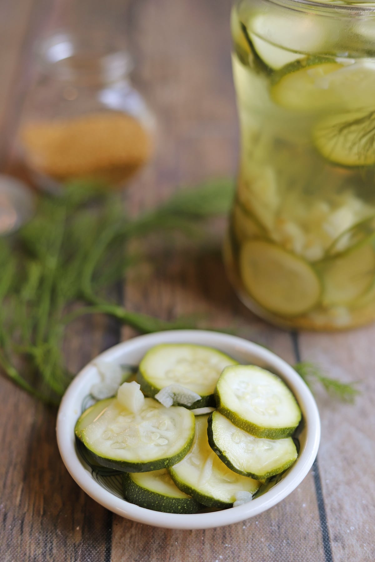 Zucchini quick pickles in glass dish by jar, dill, and mustard seeds.