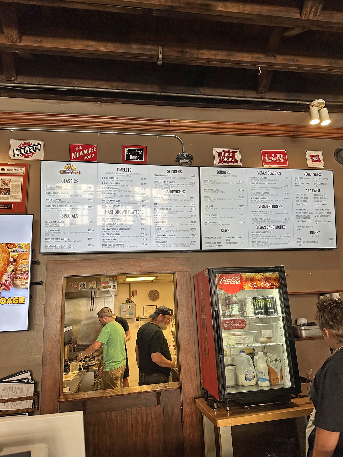 Wall menu display at Railroad Bill's Dining Car, and opening to kitchen, where people are cooking.