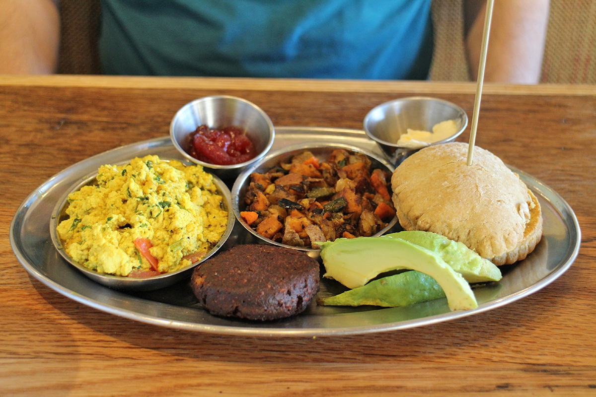 Metal plate with tofu scramble, sweet potato hash, biscuit, veggie sausage, and sliced avocado.