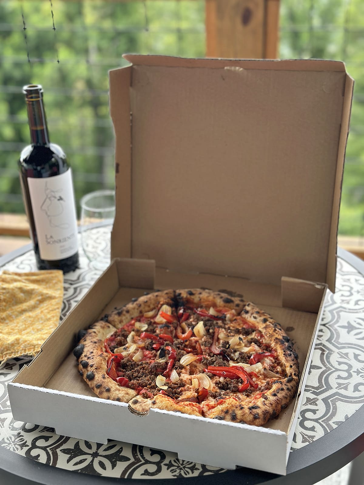 Vegan pizza in box with vegan hot Italian sausage, fire roasted onions, fire roasted red peppers, and vegan mozzarella. Also on table, La Sonriente wine and a cloth napkin.