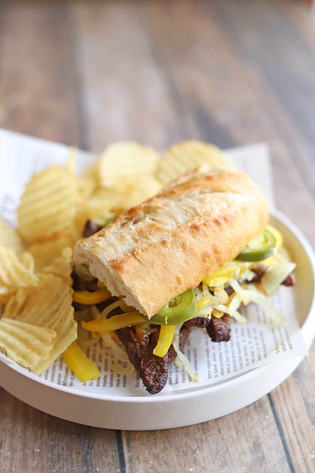 Beefless bulgogi sandwich with peppers, onions, hot peppers, and vegan cheese by side of potato chips.