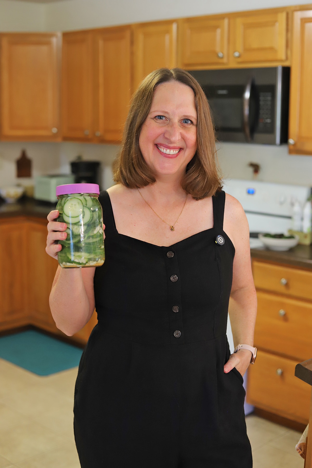 Cadry wearing a black romper and holding a jar of quick pickled cucumbers in the kitchen.