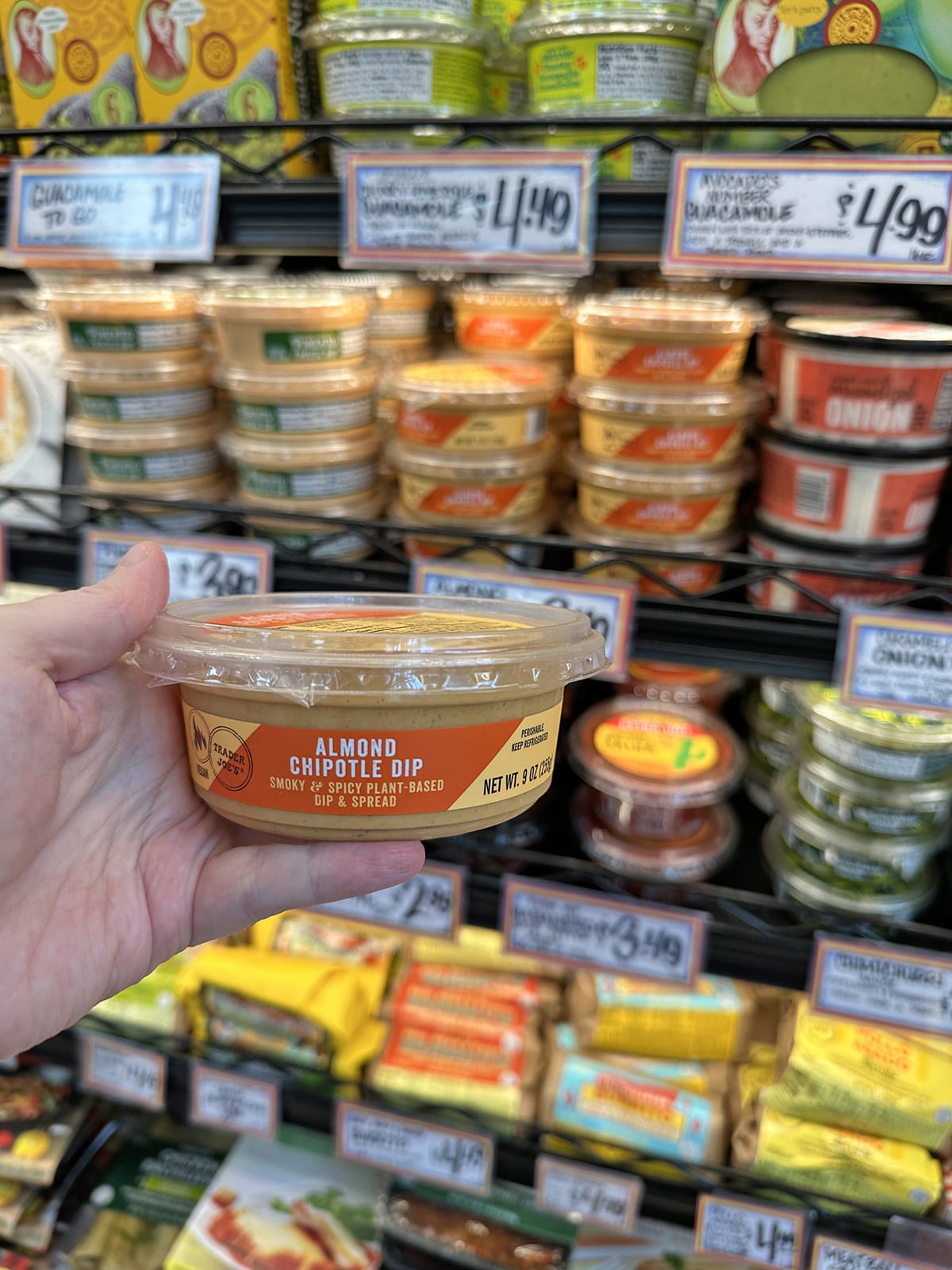 Hand holding container of almond chipotle dip in front of refrigerated case.