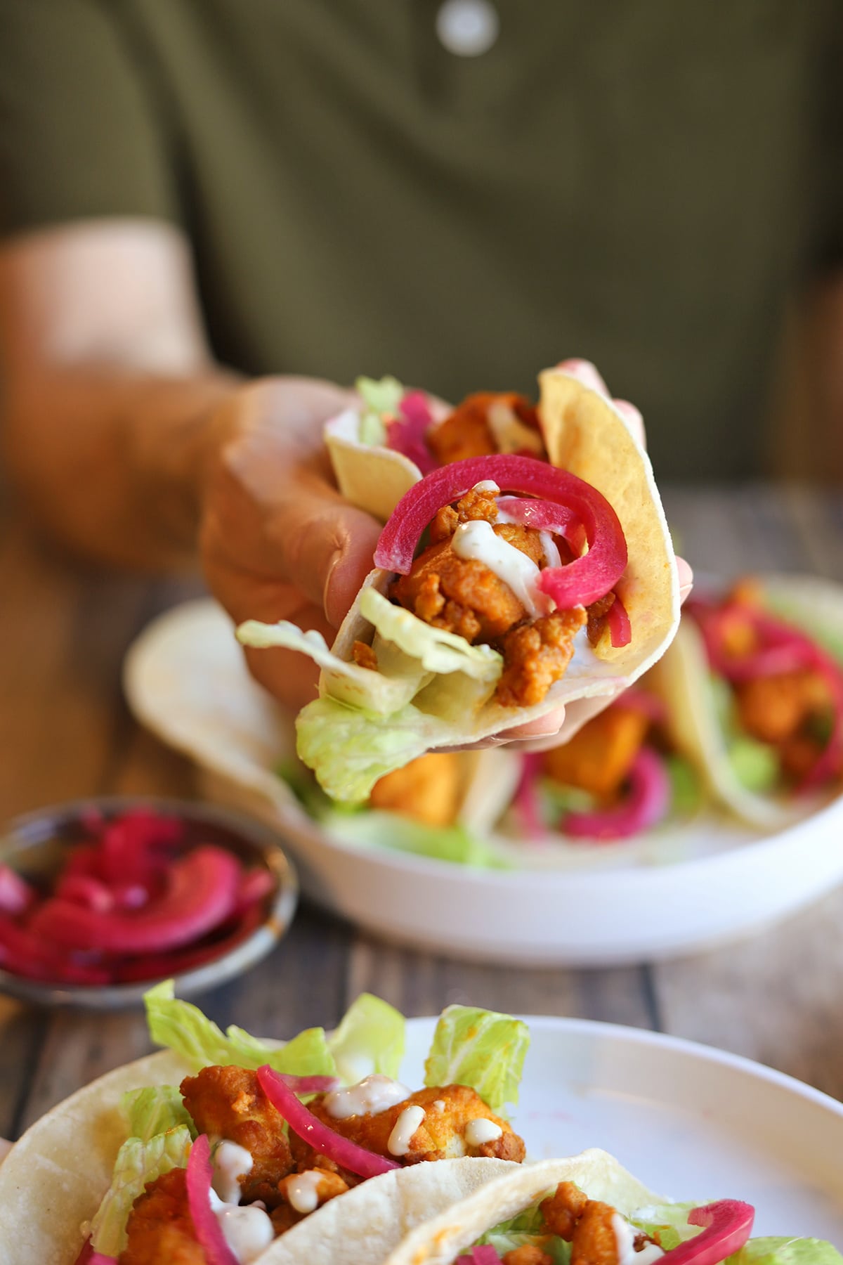 Hand holding vegan buffalo tofu tacos in corn tortilla topped with lettuce, pickled onions, and creamy dressing.