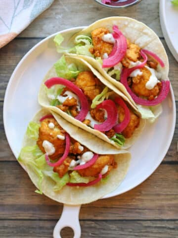 Three buffalo tofu tacos on plate with lettuce, pickled onions, and dollops of creamy dressing.