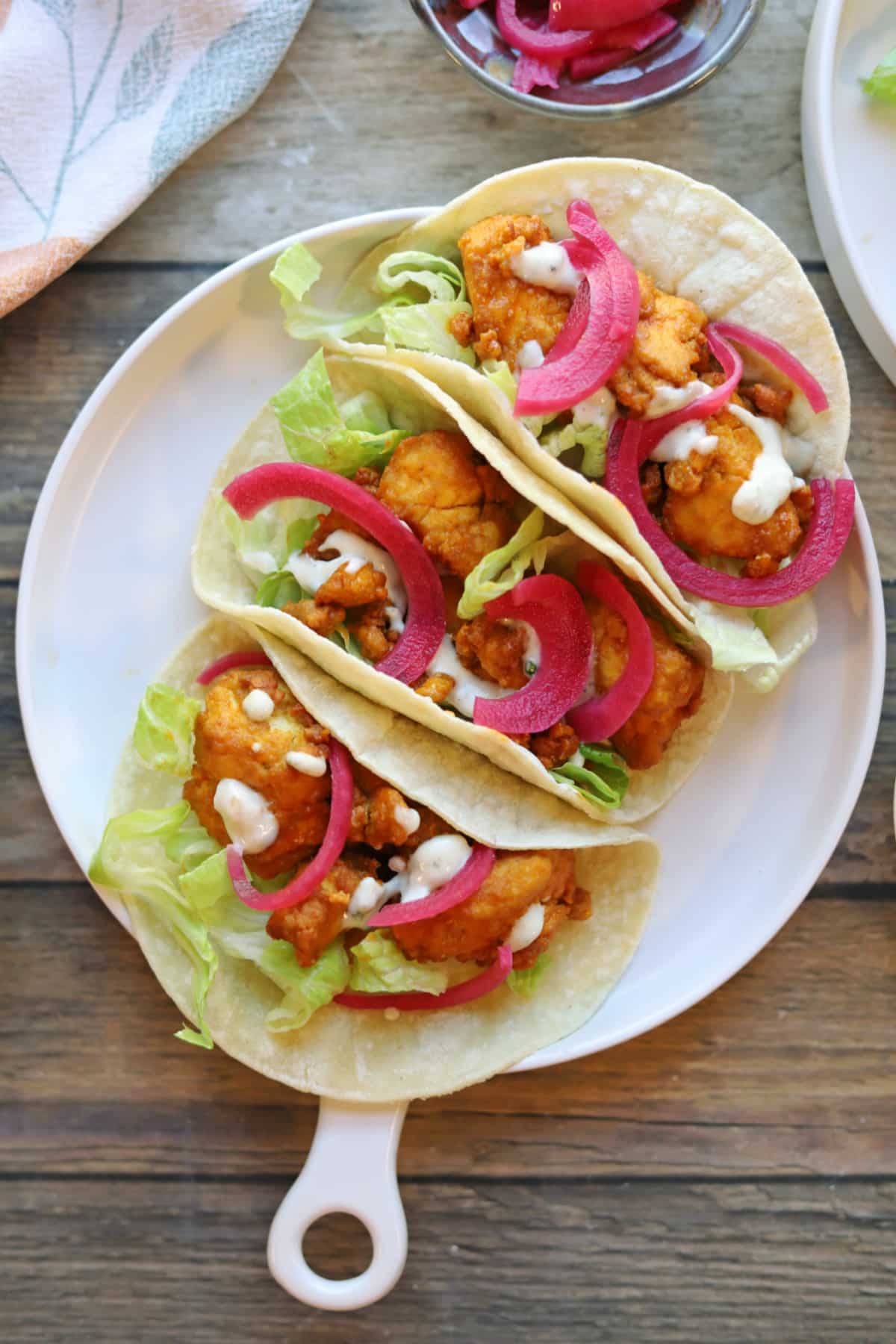 Three buffalo tofu tacos on plate with lettuce, pickled onions, and dollops of creamy dressing.