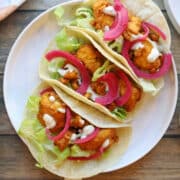Crispy buffalo tofu tacos on plate, topped with pickled red onions and creamy onion & garlic vegan dressing.