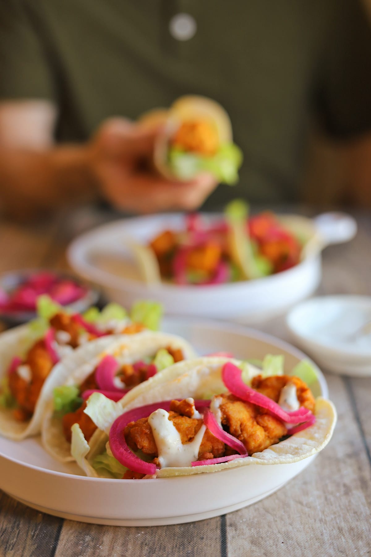 Vegan buffalo tacos with creamy onion and garlic dressing on platter, while hand holds taco in background.